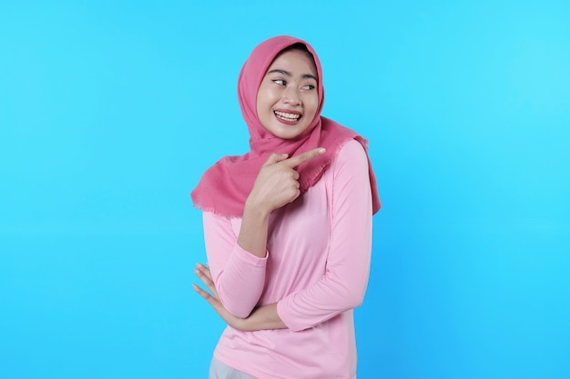 Femme asiatique souriante avec son doigt pointé isolé sur fond bleu clair portant un hijab avec un t-shirt rose