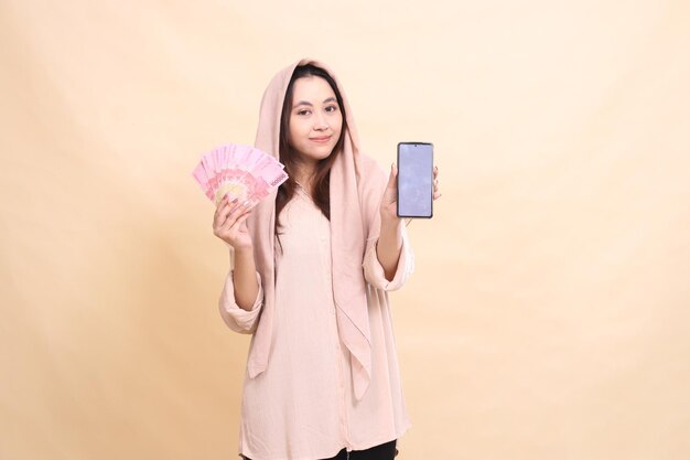 Une femme asiatique sourit heureuse à la caméra en tenant de l'argent en rupies et un écran de téléphone portable.