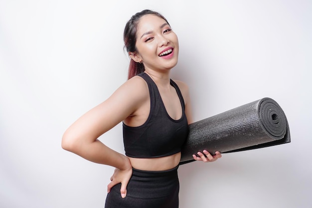 Une femme asiatique sportive souriante portant des vêtements de sport tient un tapis de yoga sur fond blanc isolé