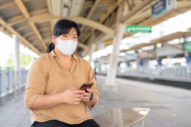 Femme asiatique en surpoids avec masque pensant tout en utilisant le téléphone à la gare du ciel