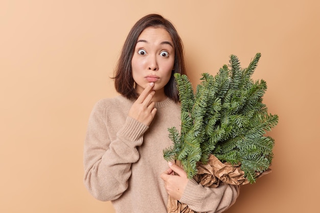 Une femme asiatique surprise et impressionnée tient des branches de sapin vert disposées en bouquet et enveloppées dans du papier regarde les yeux sur écoute se prépare pour des vacances isolées sur fond beige Concept d'heure d'hiver