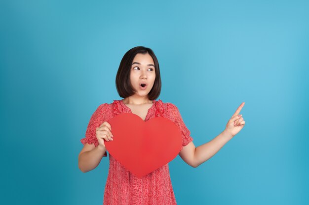 Femme asiatique surprise avec sa bouche ouverte tient un grand coeur de papier rouge et pointe avec son index sur un espace vide pour le texte