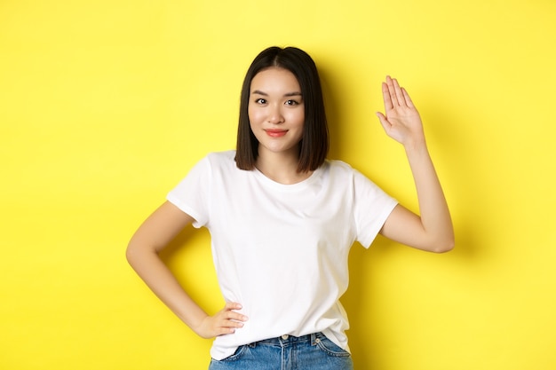 Femme asiatique sympathique en t-shirt blanc en agitant la main et en disant bonjour, en vous saluant, debout sur le jaune.