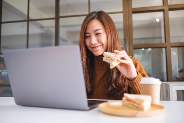 Une femme asiatique tenant et mangeant un sandwich de blé entier tout en travaillant sur un ordinateur portable