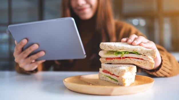 Une femme asiatique tenant et mangeant un sandwich de blé entier tout en utilisant un tablet pc