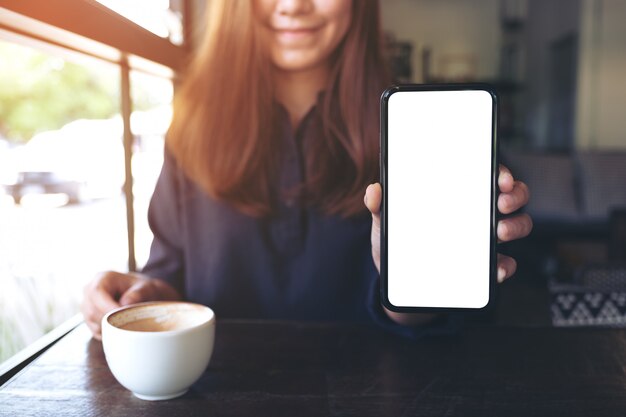 Une femme asiatique tenant et montrant un téléphone mobile noir avec écran de bureau blanc vierge avec des tasses à café sur table en bois vintage