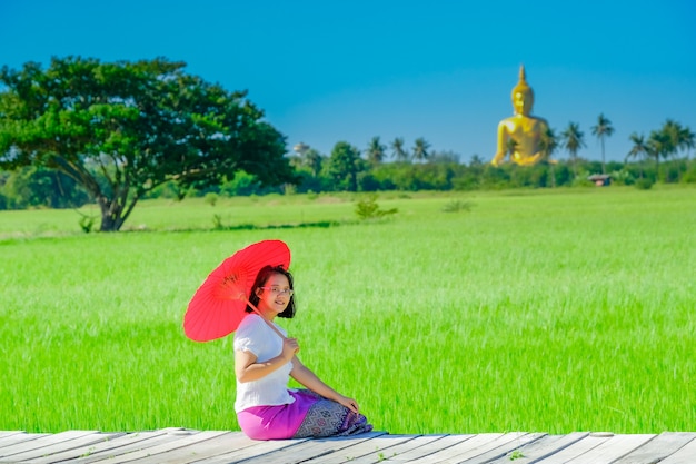 Une femme asiatique tenant un parapluie rouge assis sur un pont en bois dans une rizière avec une grande image de Bouddha doré.