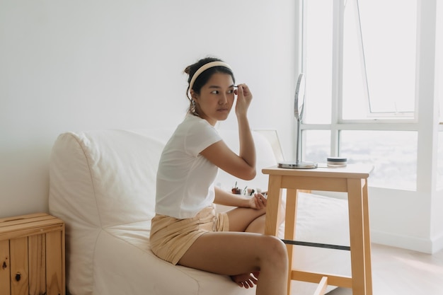 Une femme asiatique thaïlandaise utilisant un crayon et dessinant des sourcils assise sur un canapé blanc devant le miroir en