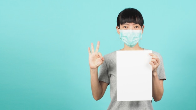 Une femme asiatique tient du papier A4 vierge et fait un signe de main OK et porte un masque facial ou un masque médical sur fond bleu et vert ou menthe.