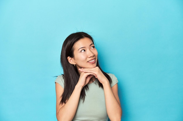 Une femme asiatique en toile de fond de studio vert d'été garde les mains sous le menton et regarde joyeusement de côté