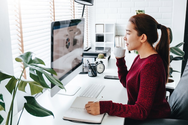 Femme asiatique travaillant à domicile utilisant un ordinateur et buvant du café dans sa chambre à coucher document financier et conférence en ligne réunion pour de nouveaux projets