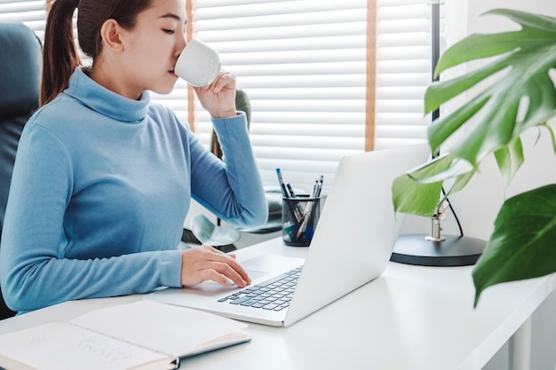 Femme asiatique travaillant à domicile utilisant un ordinateur et buvant du café dans sa chambre à coucher document financier et conférence en ligne réunion pour de nouveaux projets