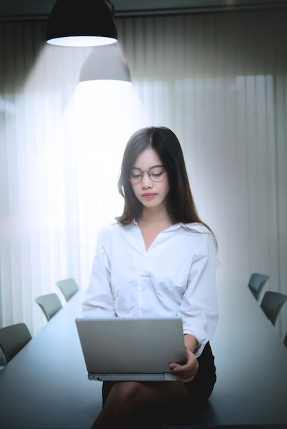 Femme asiatique travaillant des heures supplémentaires tard dans la nuit dans la lumière du bureau de la lampe pour travailler avec un ordinateur portable sur la jambe et s'asseoir au bureau