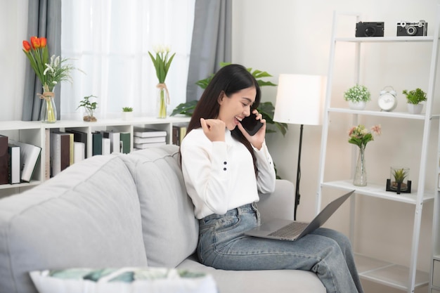 Une femme asiatique utilisant un téléphone portable avec une expression positive, un grand sourire, une robe décontractée et assise sur un canapé à fond blanc.