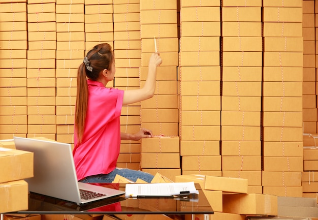 Une femme asiatique utilise un crayon pour compter les boîtes de colis dans son commerce en ligne à la maison avec un ordinateur portable sur le bureau