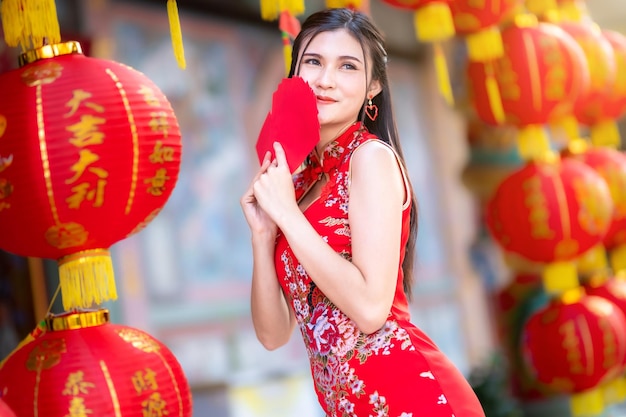 Femme asiatique vêtue d'une robe cheongsam rouge décoration traditionnelle tenant des enveloppes rouges à la main et des lanternes avec le texte chinois Bénédictions écrites dessus Est une bénédiction de fortune pour le Nouvel An chinois