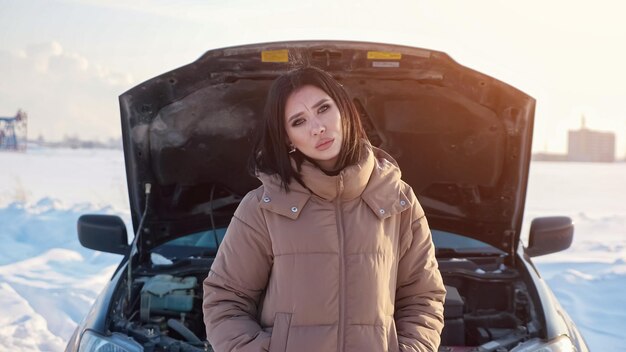 Une femme assez contrariée en veste chaude hausse les épaules debout près d'une automobile cassée avec un capot ouvert sur une route enneigée à la campagne par une froide journée d'hiver, la lumière du soleil