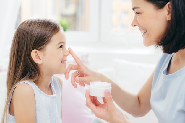 Une femme assez gaie reçoit une procédure de beauté d'une mère affectueuse. Belle jeune femme applique une crème de beauté sur le nez de sa fille