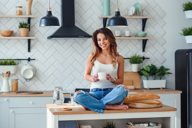 Femme assez heureuse boit du café tout en étant assis sur la table dans la cuisine confortable le matin à la maison. Détente matinale à la maison.