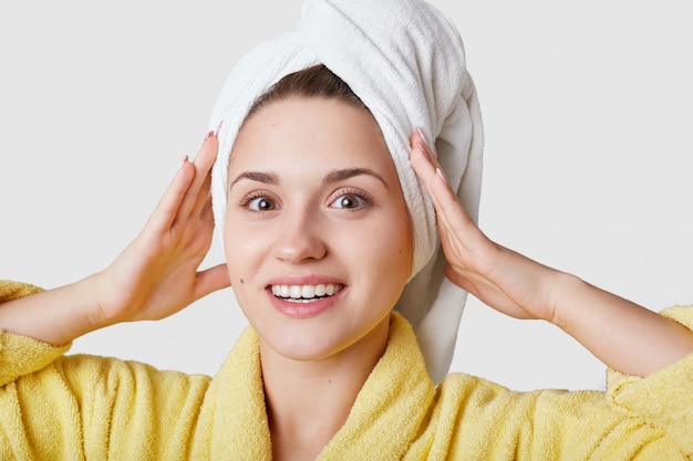 Une femme assez insouciante fait preuve de beauté naturelle, sourit largement, a la peau fraîche, garde les mains sur la tête, porte une serviette