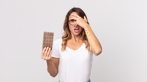 Femme assez mince à l'air choquée, effrayée ou terrifiée, couvrant le visage avec la main et tenant une barre de chocolat