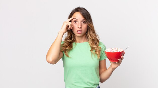 Femme assez mince ayant l'air surprise, réalisant une nouvelle pensée, idée ou concept et tenant un bol de petit-déjeuner