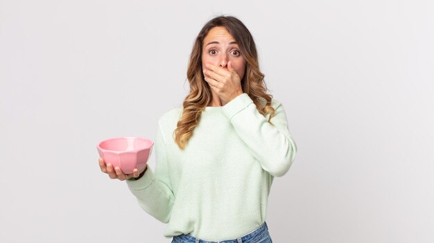 Femme assez mince couvrant la bouche avec les mains avec un choqué et tenant un bol de pot vide