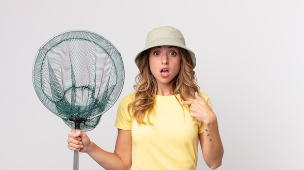 Femme assez mince qui a l'air choquée et surprise avec la bouche grande ouverte, pointant vers l'autoportant un chapeau et tenant un filet de pêche