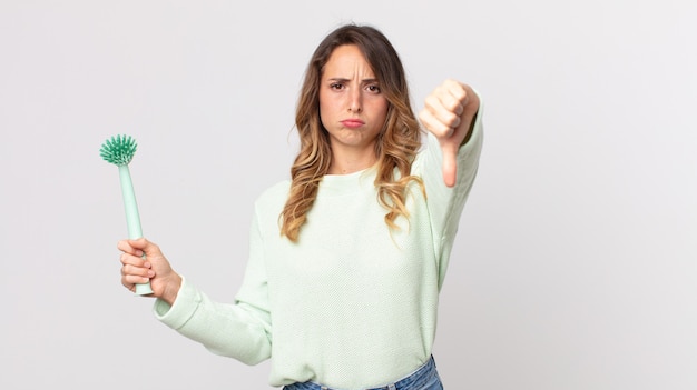 Femme assez mince se sentant croisée, montrant les pouces vers le bas et tenant une brosse à vaisselle