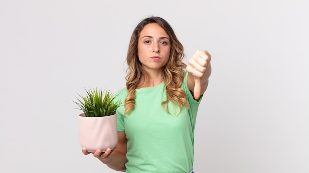 Femme assez mince se sentant croisée, montrant les pouces vers le bas et tenant une plante décorative