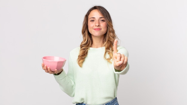 Femme assez mince souriant fièrement et avec confiance faisant le numéro un et tenant un bol de pot vide