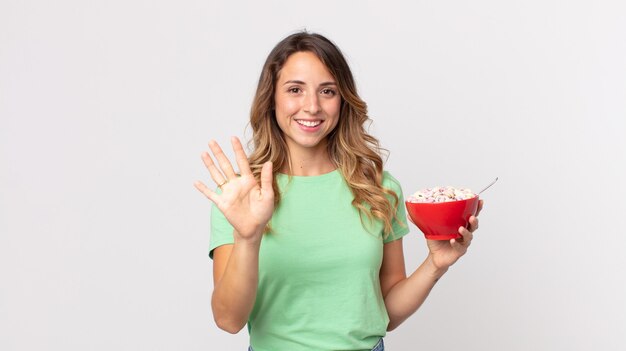 Femme assez mince souriant joyeusement, agitant la main, vous accueillant et vous saluant et tenant un bol de petit-déjeuner