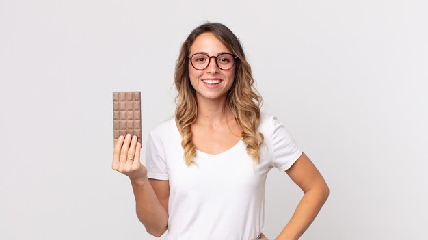 femme assez mince souriant joyeusement avec une main sur la hanche et confiante et tenant une barre de chocolat