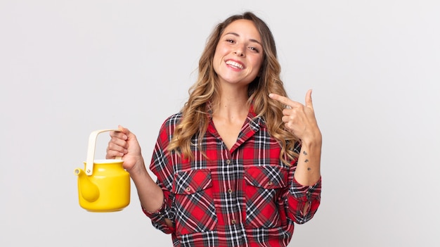 Femme assez mince souriante pointant avec confiance vers son propre large sourire et tenant une théière