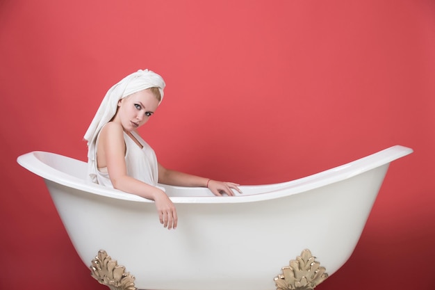 Femme assez sexy avec une serviette turban sur la tête assise dans une baignoire blanche sur fond rouge spa et beaut...