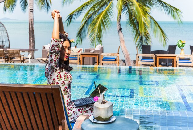 Femme assise au bord de la piscine avec vue sur l'océan travaillant sur un ordinateur portable