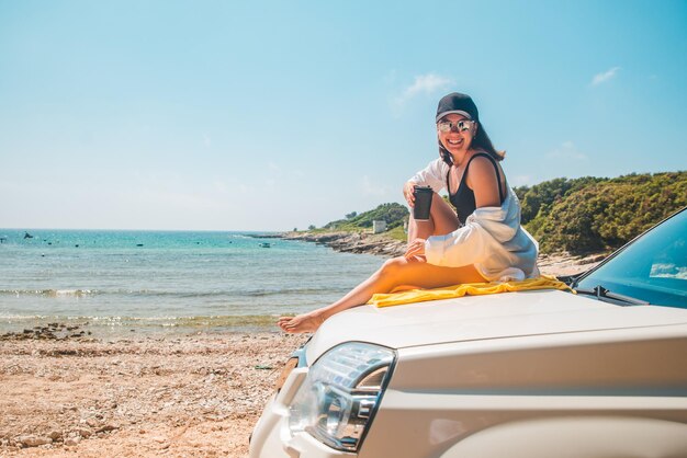 Femme assise au capot de la voiture buvant du café en profitant de la vue sur les vacances d'été sur la mer