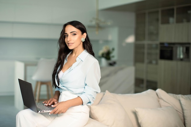 Femme assise sur un canapé à l'intérieur d'un appartement à la maison à l'aide d'un ordinateur portable