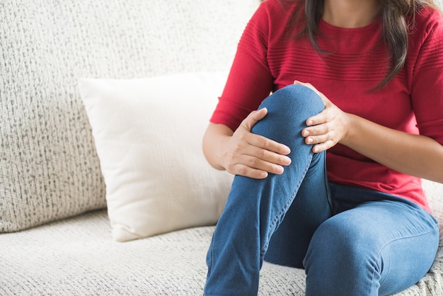 Femme assise sur le canapé et le sentiment de douleur au genou, soins de santé et concept médical.