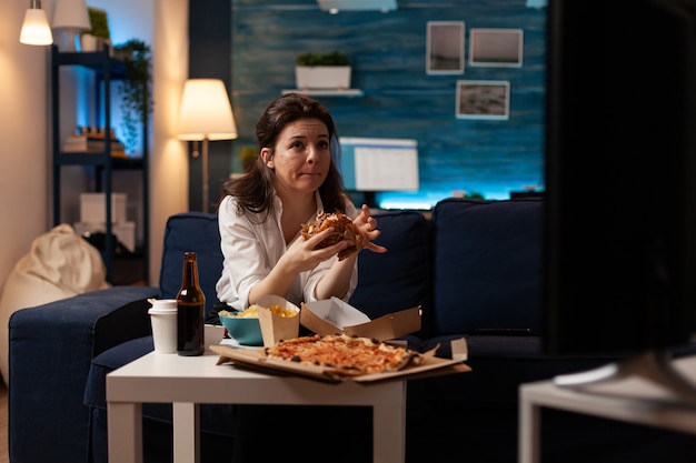 Femme assise sur un canapé en train de manger un délicieux hamburger délicieux en regardant un film documentaire