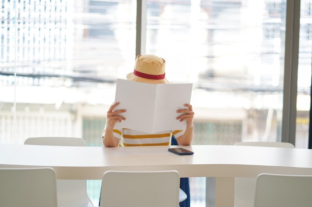 Femme assise sur une chaise et lisant un livre blanc ou un magazine dans un espace de travail ou une bibliothèque.