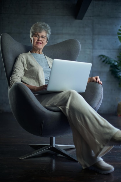 Femme assise sur une chaise moderne près de la fenêtre dans une pièce confortable et lumineuse à la maison Elle travaille sur un ordinateur portable dans une atmosphère relaxante