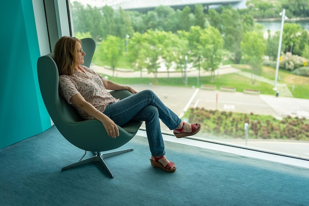 Femme assise dans un fauteuil décontracté et contemplant le paysage depuis une grande fenêtre de la pièce