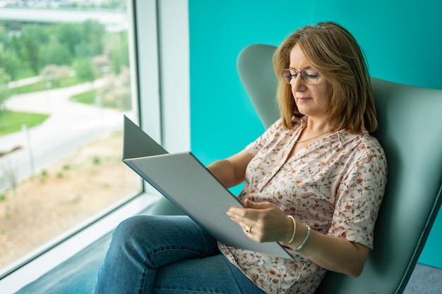 Femme assise dans un fauteuil relaxant et lisant un livre à côté d'une immense fenêtre panoramique de la pièce