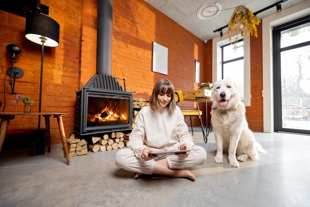 Femme assise détendue avec son chien sur le sol près de la cheminée et utilisant une tablette numérique à la maison