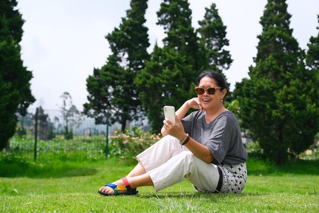 Une femme assise sur l'herbe tenant un smartphone effectuant un appel vidéo dans le parc