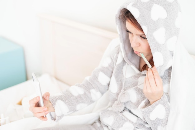 Femme assise sur le lit recouvert d'une couverture et photographiée au téléphone avec un thermomètre dans la bouche Une fille enrhumée prend un selfie sur son blog Mesure de température élevée