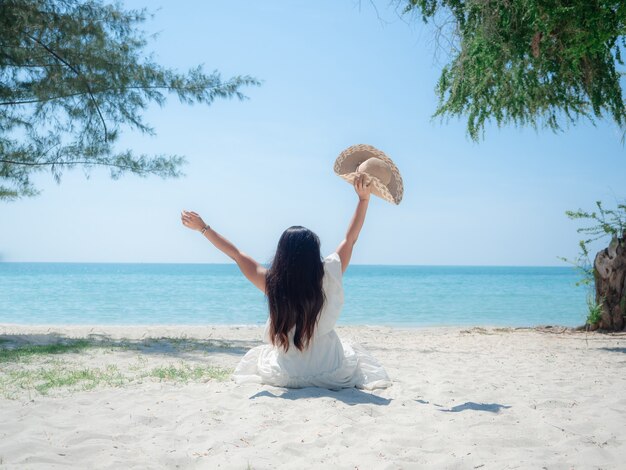 Femme assise sur la plage à la plage montre la main et chapeau joyeux sur concept de vacances et arbre