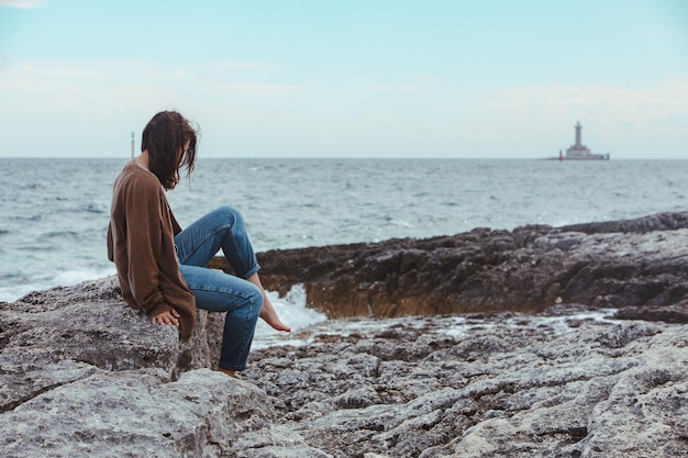 Femme assise près de la plage de la mer rocheuse dans le phare de jeans mouillés sur fond