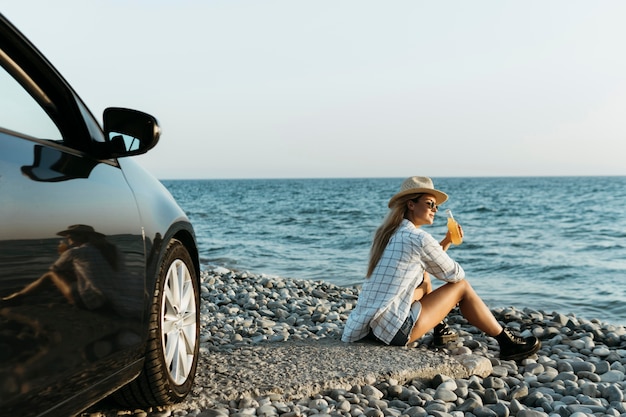 Femme assise sur des rochers avec du jus en regardant la mer près de voiture